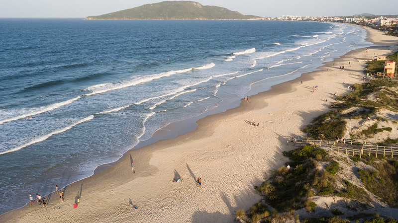 Vista aérea de uma praia extensa com areia clara e mar azul com pequenas ondas; pessoas caminham e se exercitam na faixa de areia. Ao fundo, uma ilha e uma cidade costeira são visíveis ao longo do litoral.