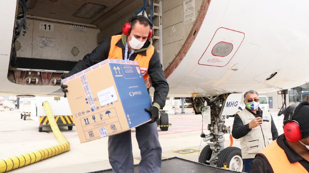 Homem de colete laranja e camisa preta retirando caixas de vacina do avião branco