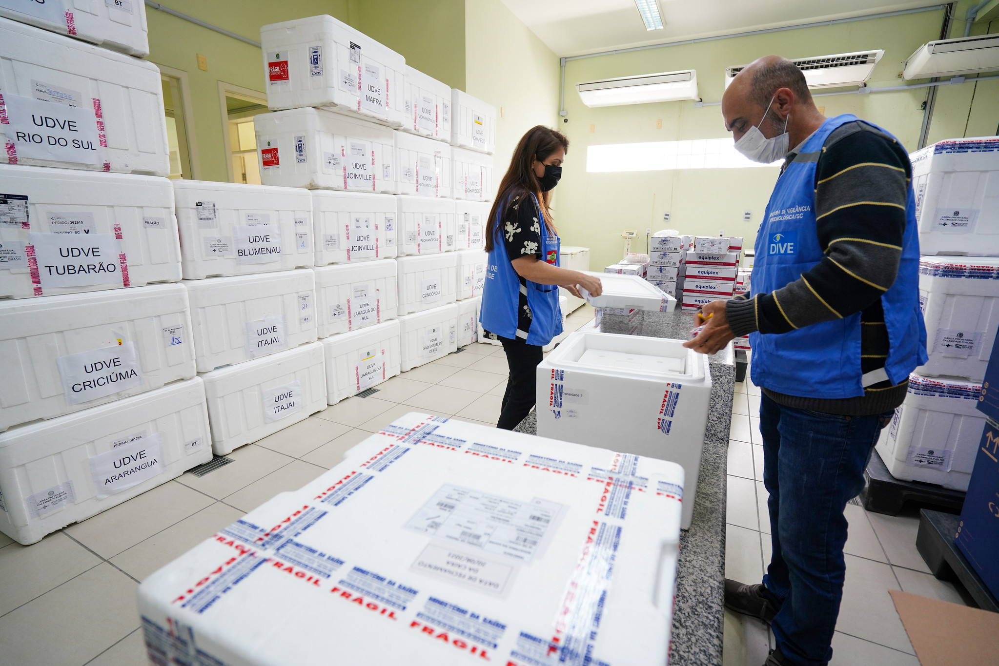 Duas pessoas com roupa preta e colete azul, um homem e uma mulher, dentro de uma sala com várias caixas térmicas