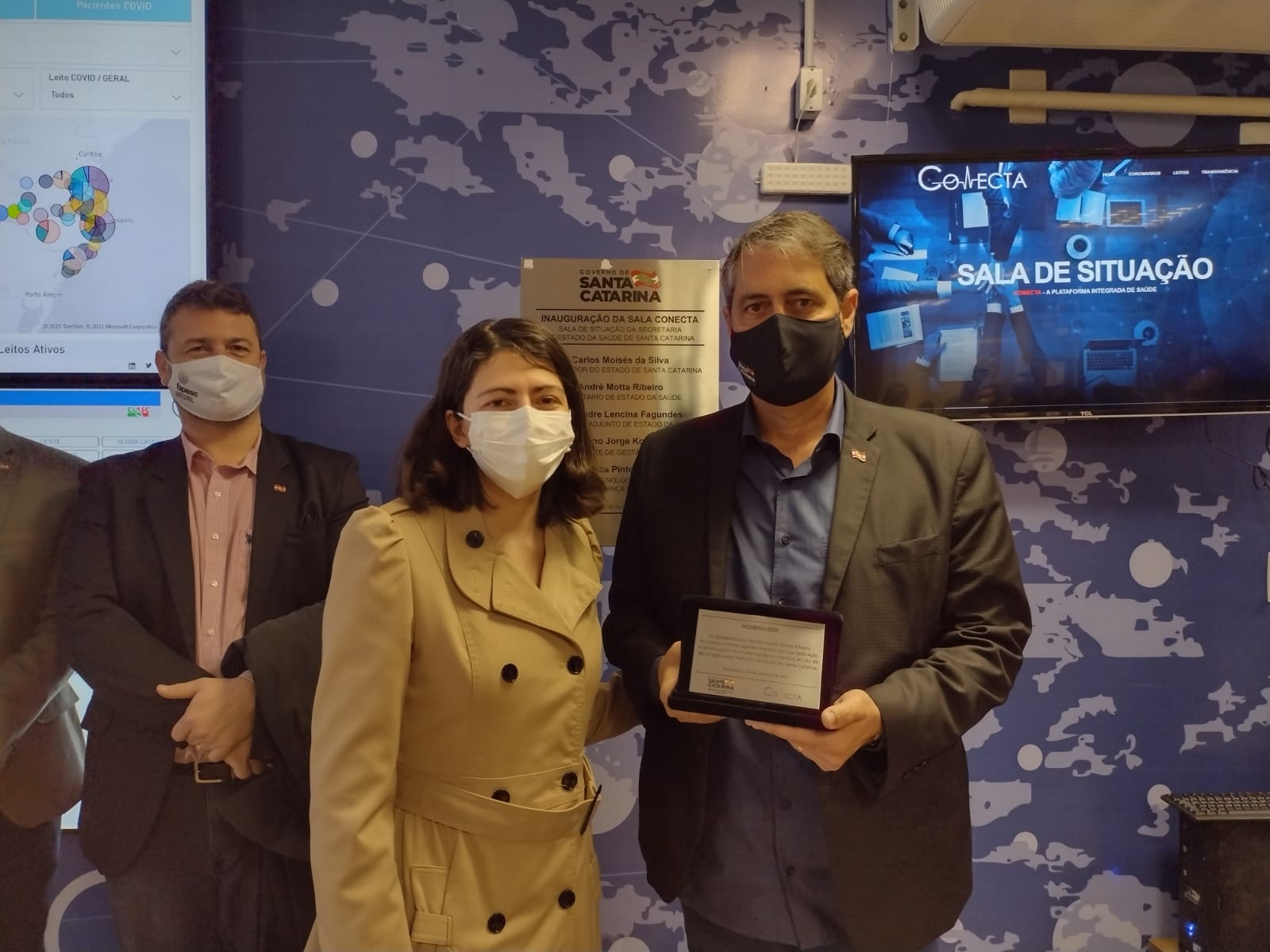 Três pessoas usando máscaras posam para foto em ambiente interno com parede azul decorada com mapa e telas ao fundo. Uma mulher de casaco claro está ao lado de um homem de terno escuro que segura uma placa de homenagem. No fundo, há uma tela exibindo a frase 'SALA DE SITUAÇÃO' e um painel do Governo de Santa Catarina.