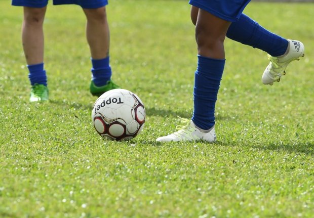 Duas crianças usando uniformes azuis e chuteiras, uma delas prestes a chutar uma bola de futebol branca com detalhes vermelhos e a marca Topper, em um campo de grama verde.