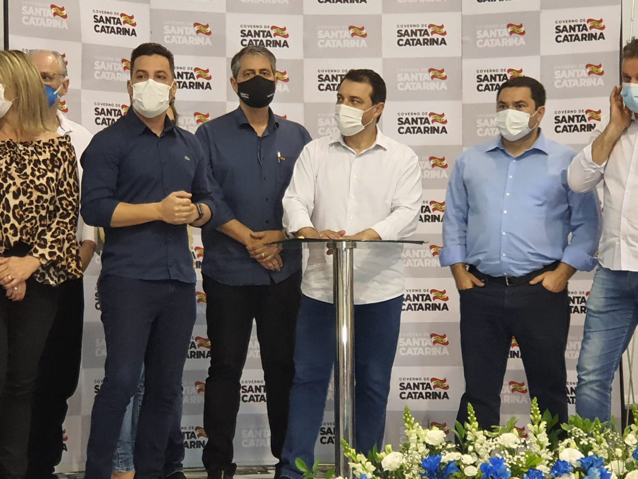 Grupo de pessoas em pé, todas usando máscaras, durante um evento oficial do Governo de Santa Catarina. Ao centro, um homem de camisa branca e calça azul fala ao microfone, atrás de um púlpito de metal. Ao fundo, há um painel com o logotipo do governo estadual repetido. À frente, arranjos de flores brancas e azuis decoram o espaço. Os participantes vestem roupas sociais e demonstram atenção ao momento.