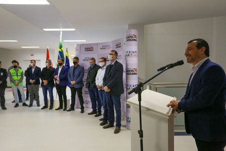 Homem de terno fala sorrindo ao microfone em um púlpito, enquanto um grupo de pessoas, todas de máscara, está alinhado ao fundo em frente a painéis com o logotipo do Governo de Santa Catarina e bandeiras do Brasil e do estado. O ambiente é claro e moderno, sugerindo um evento oficial ou inauguração.