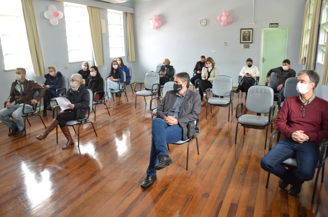 O secretário de Estado da Saúde, André Motta Ribeiro, sentado em sala da Maternidade Carmela com várias outras pessoas. A sala possui piso de madeira, paredes cinzas e tem cadeiras cinzas.