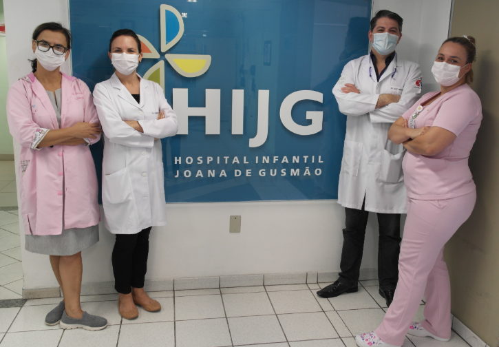 Equipe médica do Hospital Infantil composta por duas mulheres à esquerda e um homem e uma mulher à direita, todos usando máscaras cirúrgicas, durante atendimento a crianças com deformidades craniofaciais congênitas.