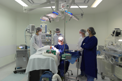 Equipe médica composta por cinco profissionais, todos usando máscaras, toucas e roupas cirúrgicas, realiza um procedimento em uma sala de cirurgia bem iluminada. O cirurgião está sentado operando, enquanto os demais acompanham e auxiliam. Ao lado, há equipamentos médicos e uma incubadora neonatal.