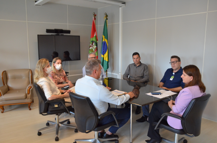 Sete pessoas sentadas ao redor de uma mesa de reuniões em um ambiente institucional. Algumas usam máscaras faciais. Ao fundo, as bandeiras do Brasil e de Santa Catarina estão posicionadas ao lado de uma televisão. Os participantes aparentam estar atentos e envolvidos na discussão.