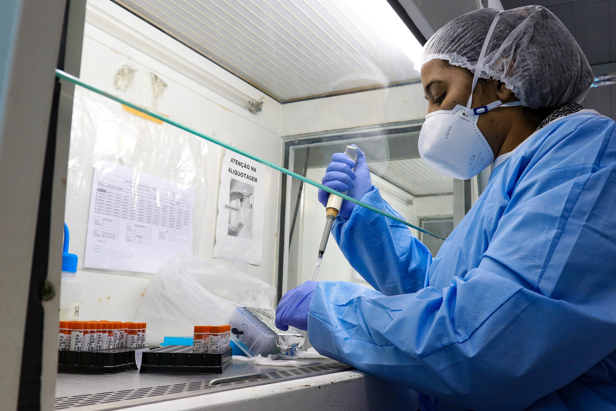 Profissional do Laboratório Central de Saúde Pública (LACEN/SC) manuseia micropipeta em cabine de segurança biológica, usando equipamentos de proteção como máscara PFF2, touca, avental e luvas. À frente, tubos de ensaio identificados e caixas criogênicas organizadas sobre a bancada.