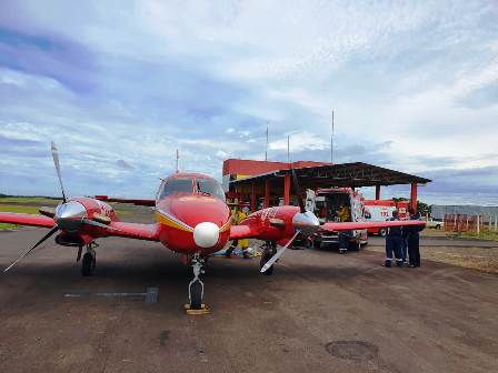 Avião vermelho do Corpo de Bombeiros estacionado em pista de aeródromo com equipe médica e bombeiros ao redor, próximo a uma ambulância e estrutura coberta ao fundo, sob céu parcialmente nublado.