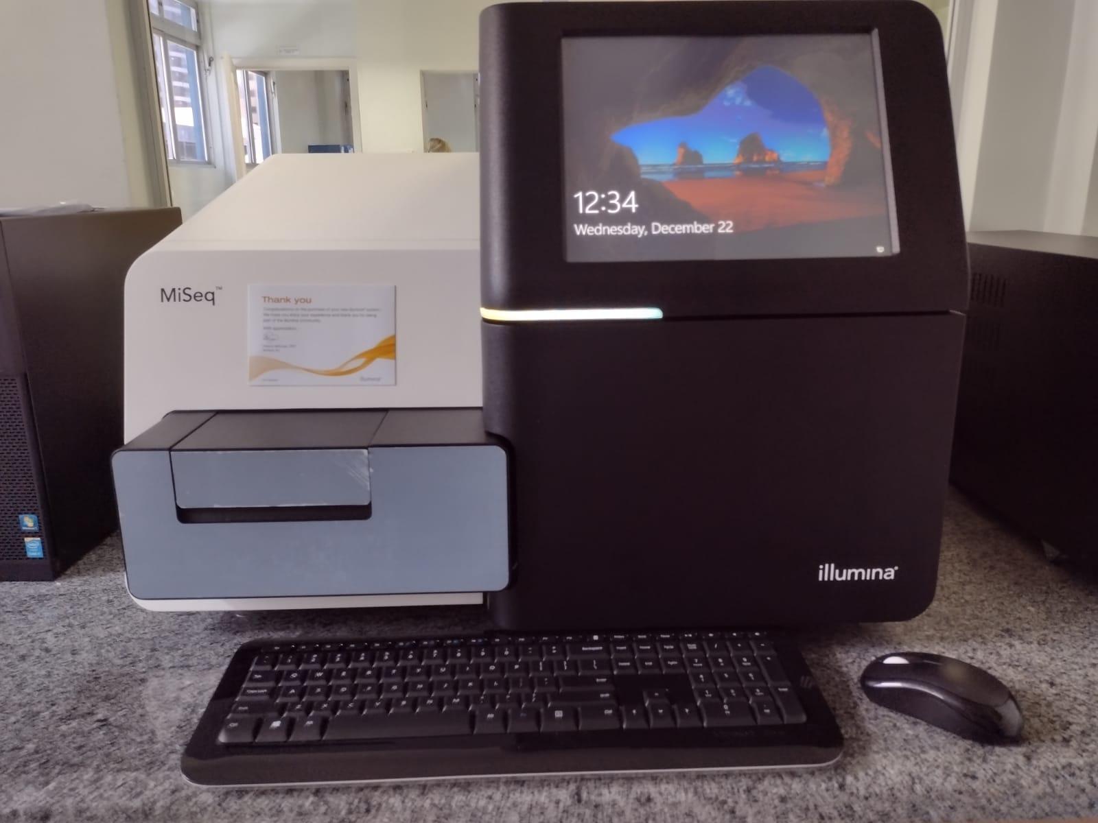 Sequenciador MiSeq da Illumina em bancada de laboratório, com teclado e mouse posicionados à frente e tela exibindo a data e hora.