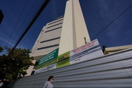 Prédio alto de vários andares visto de baixo para cima, com fachada clara e moderna. Na frente, há um tapume metálico com placas do Governo de Santa Catarina informando sobre a construção do Centro de Hematologia e Hemoterapia. Uma pessoa usando máscara caminha na calçada em frente ao tapume, sob céu azul e fios de energia.