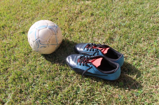 Bola de futebol branca com detalhes azuis ao lado de um par de chuteiras pretas e azuis com cadarços pretos, posicionados sobre a grama verde sob luz do sol.