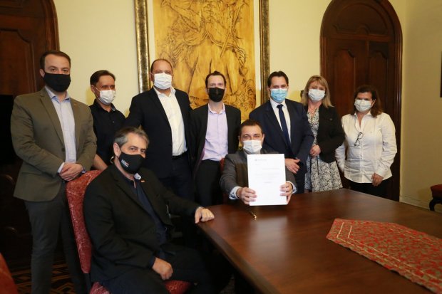 Grupo de nove pessoas posando para foto em uma sala de reuniões com móveis de madeira escura e tapeçaria clássica. O ambiente tem portas de madeira e uma grande obra de arte dourada na parede ao fundo. Todos estão usando máscaras faciais, indicando medidas de proteção contra a COVID-19. No centro da imagem, um homem sentado segura um documento assinado, enquanto as demais pessoas — em pé e sentadas — o cercam, sugerindo um momento de sanção ou assinatura oficial de uma lei ou decreto. A atmosfera é formal e institucional.