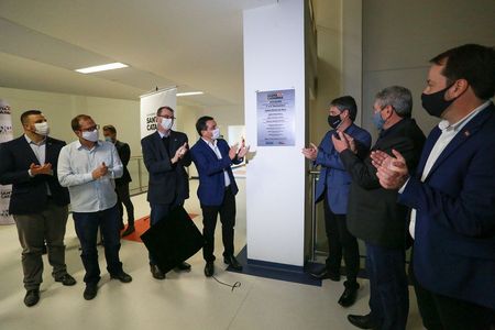 Grupo de homens usando máscaras aplaude ao redor de uma placa de inauguração fixada em uma coluna branca, com o logotipo do Governo de Santa Catarina. O ambiente é claro e moderno, sugerindo uma cerimônia oficial de inauguração.