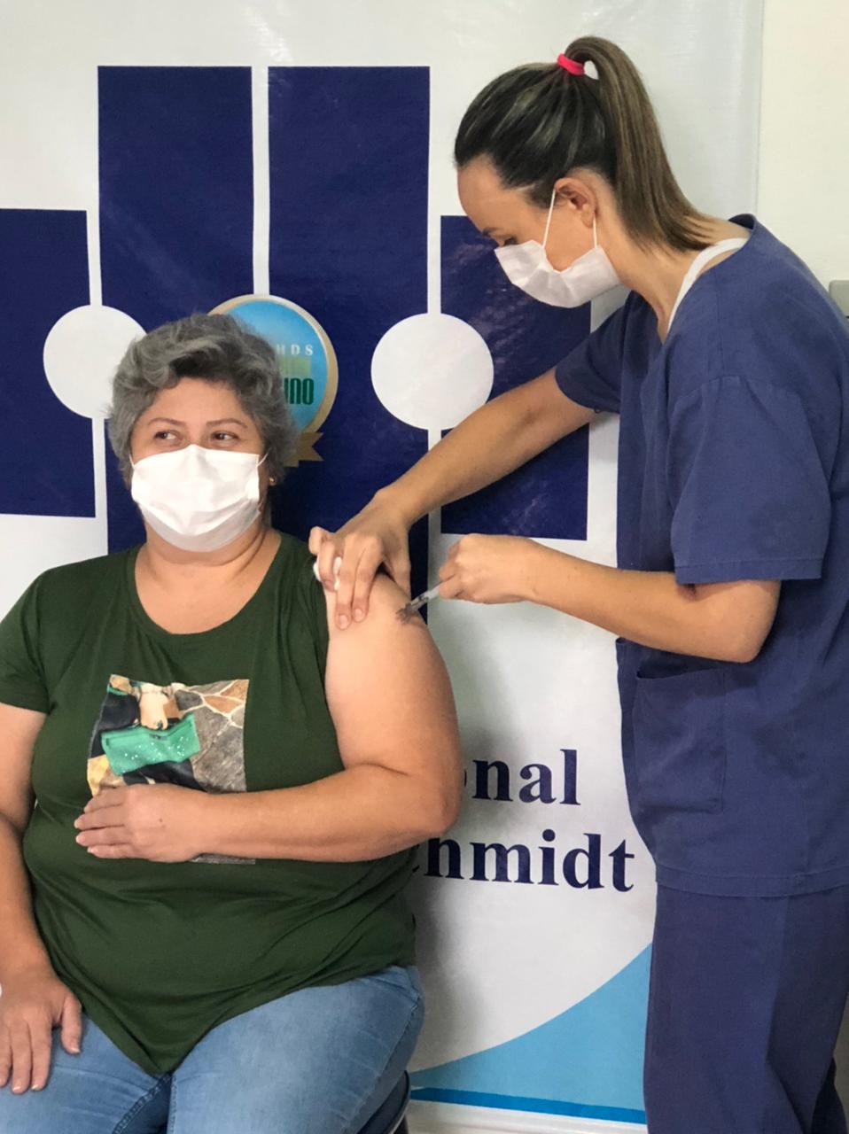 Mulher de camiseta verde e máscara branca recebe vacina no braço esquerdo aplicada por profissional de saúde com roupa azul e máscara. Ambas estão em frente a um painel branco e azul com logotipo e texto parcialmente visível. A mulher vacinada está sentada e olha para a aplicadora com expressão tranquila.